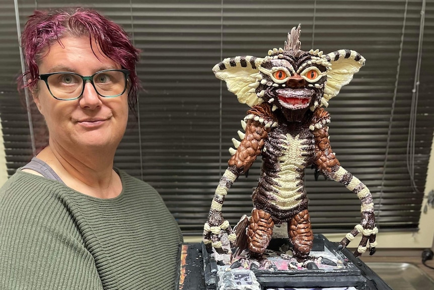 A woman smiles at the camera holding a platform that has a gremlin cake on it