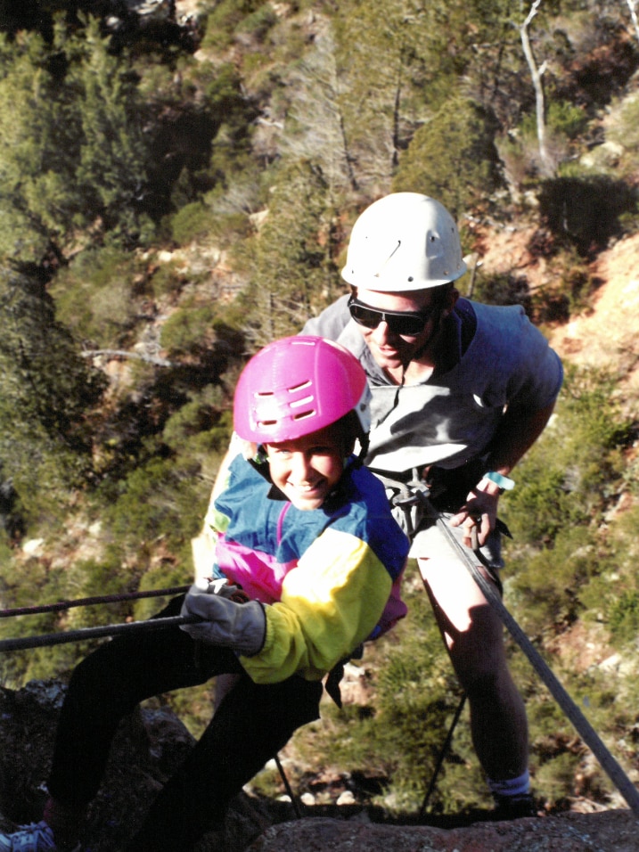 A child smiles as she climbs a rope, as an adult man follows closely behind.
