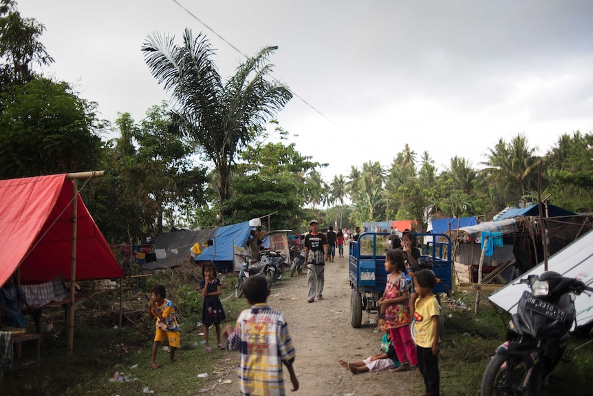 A man walks on a road with tents and palm trees in the background and children standing around.