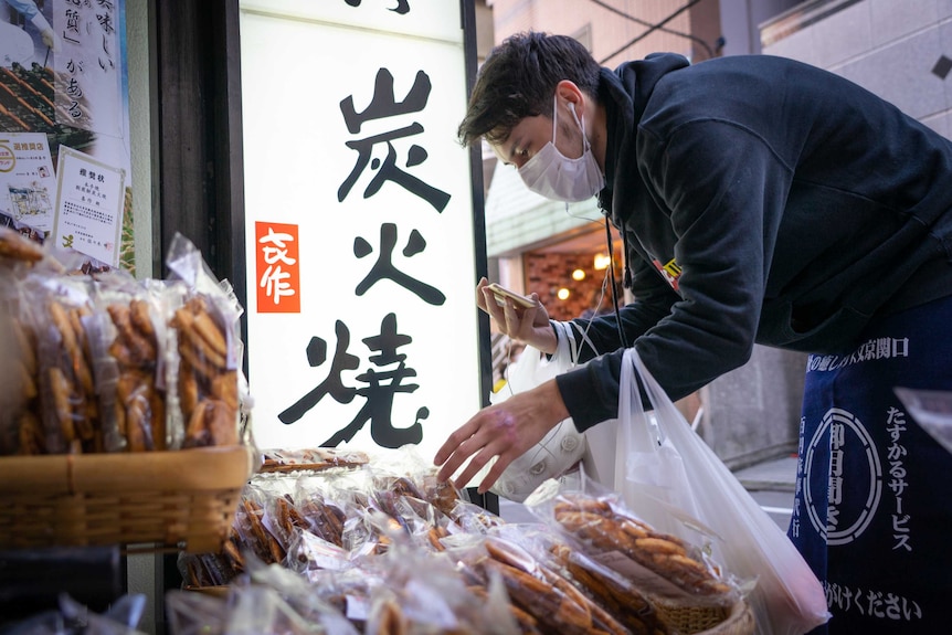 A person with black hair and wearing a hoodie and a mask leans down to pick up some groceries.