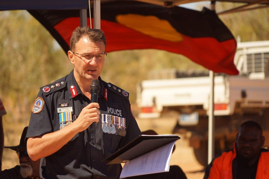 A man in police uniform standing in front of an Aboriginal flag reading a speech