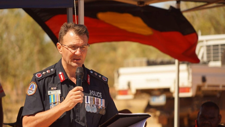 A man in police uniform standing in front of an Aboriginal flag reading a speech