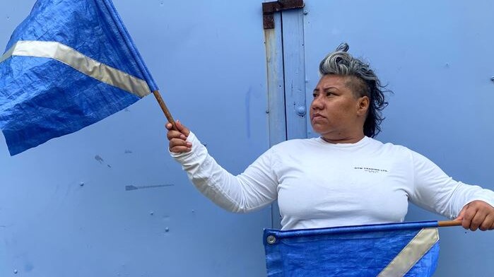 Photo of Latai Taumoepeau looking to the side, standing in front of a blue background and waving blue flags