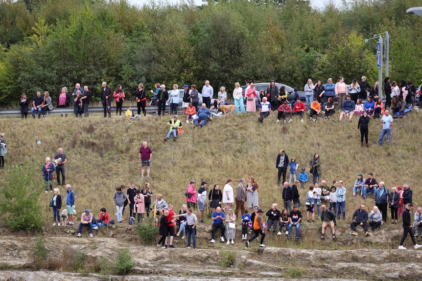 People sit and stand on a steep hill along a motorway. 
