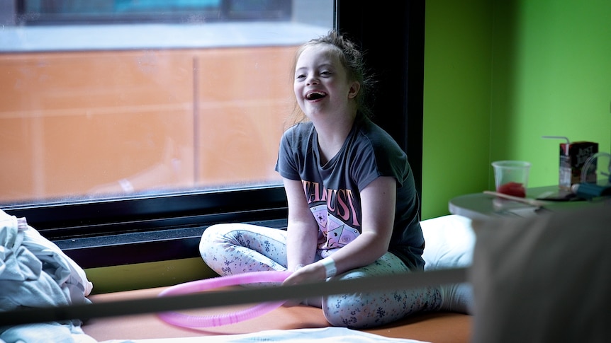 Olivia, a young girl, laughs, holding a balloon and sitting on a hospital bed.