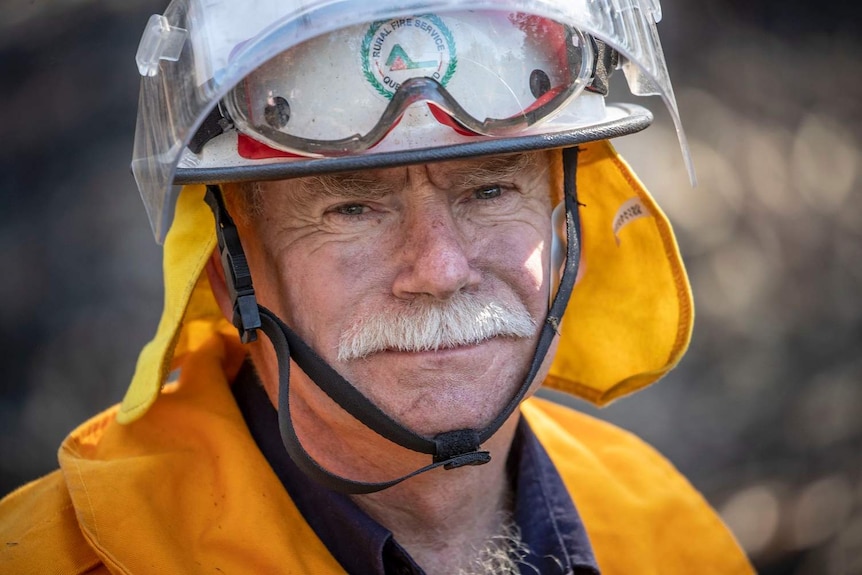 Fire fighter Peter Orth stares at camera with helmet on, wearing orange protective clothing.