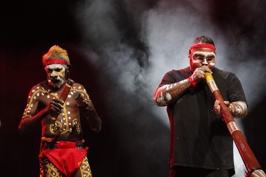 Two Indigenous men stand on a smoky stage, one playing a didgeridoo and the other dressed in traditional dress 