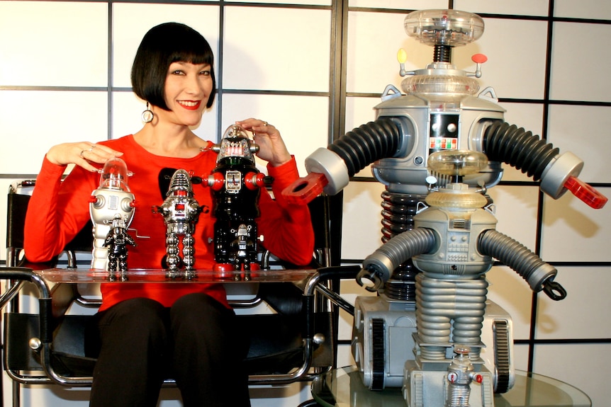 A smiling woman with an angular bob haircut posing with a collection of robot toys.