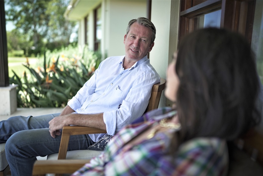 A man and woman sit on outdoor furniture looking at each other, man in focus