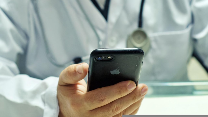 A doctor wearing a stethoscope and scrubs holds a phone.