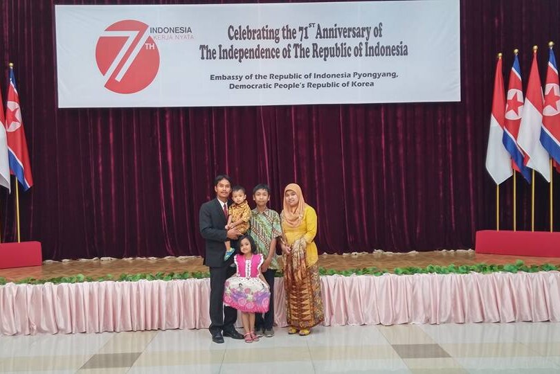 A family of 5 stands in front of a sign saying celebrating the Indonesian embassy in Pyongyang, surrounded by flags
