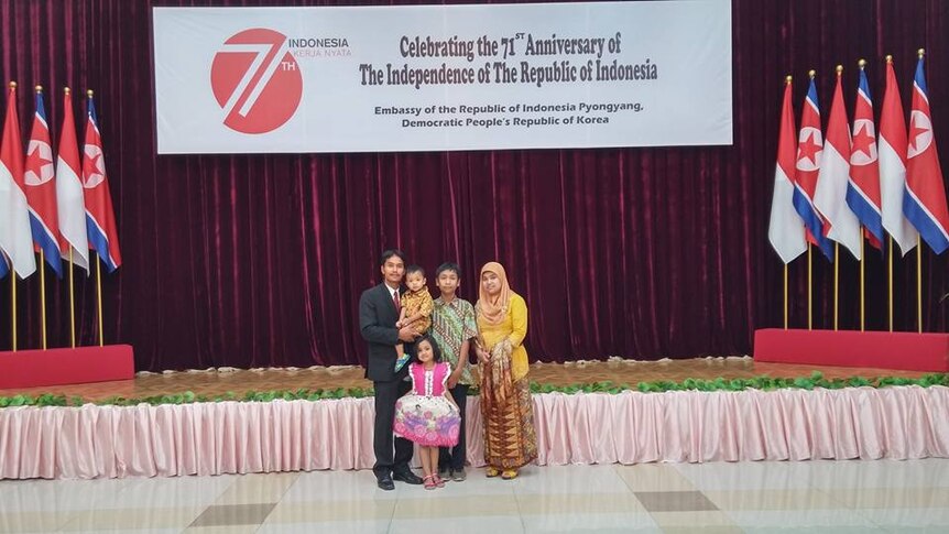 A family of 5 stands in front of a sign saying celebrating the Indonesian embassy in Pyongyang, surrounded by flags