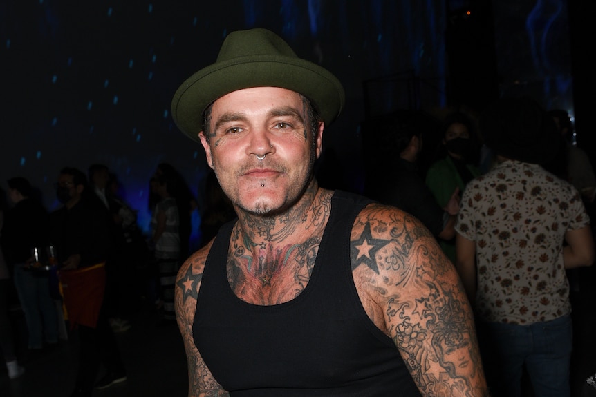 A man with tattoos on his arms, chest and neck poses for a photo, wearing a black tank top and a hat