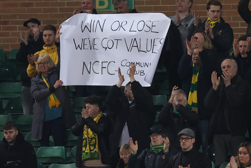 Fans in the stand at a Premier League match hold up a banner saying 'Win or Lose, We've Got Values - NCFC Community Club'.