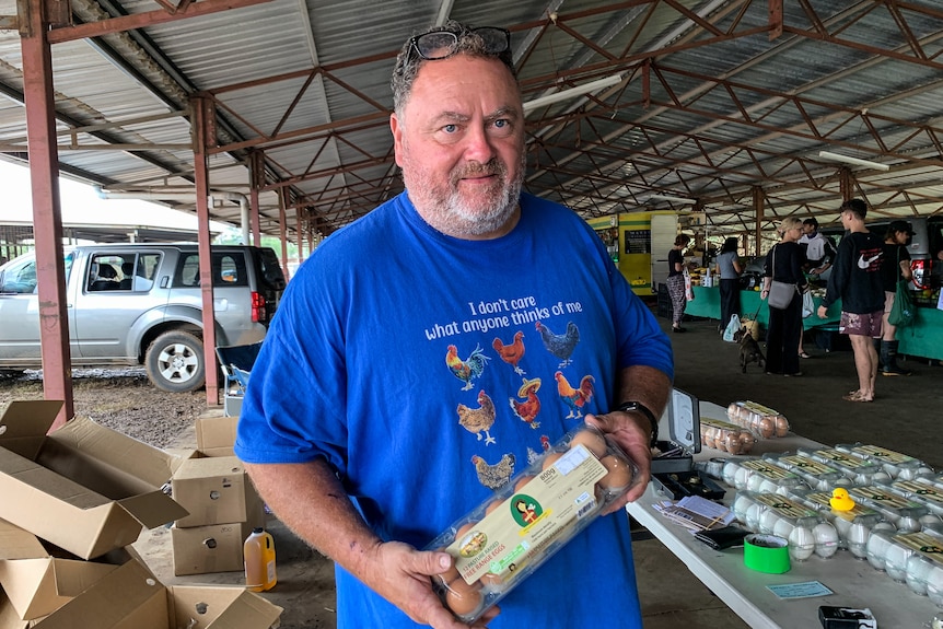 A man in a blue shirt stands holding a carton of eggs.
