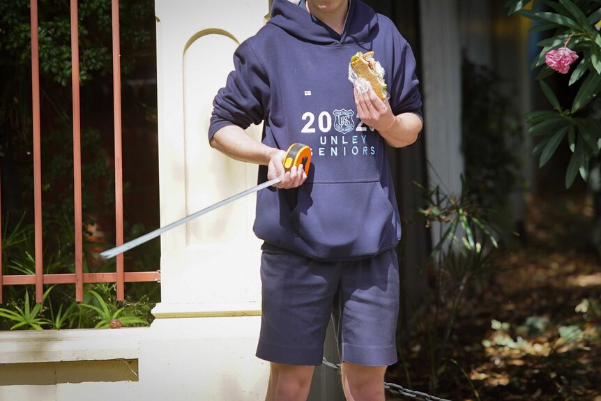 A boy wearing a jumper with 2020 Unley Seniors on it and holding a measuring tape and a sandwich
