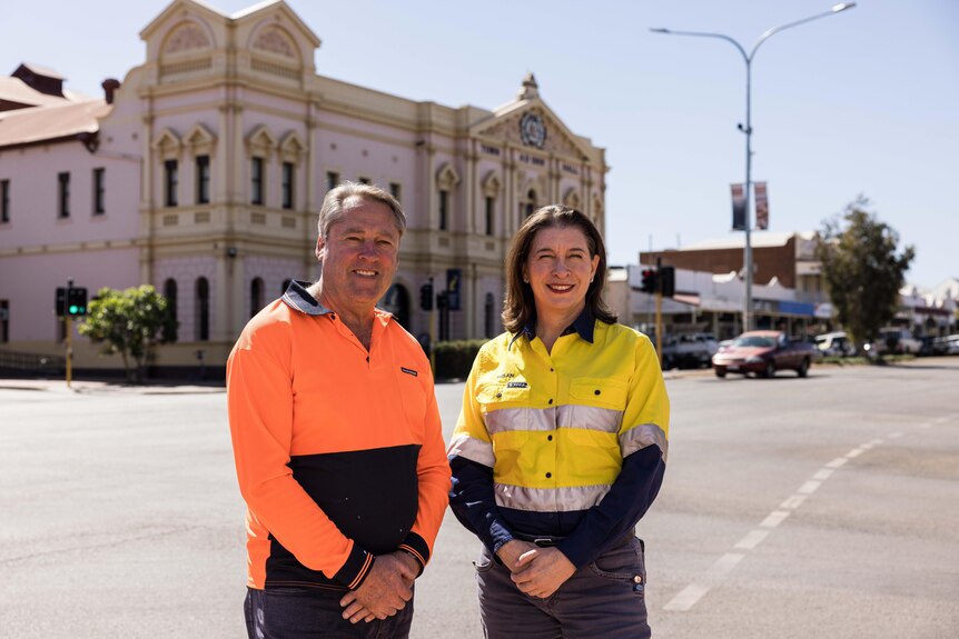 A smiling man and a woman wearing high-vis clothing, a two-storey heritage building behind them, road, cars, shops.