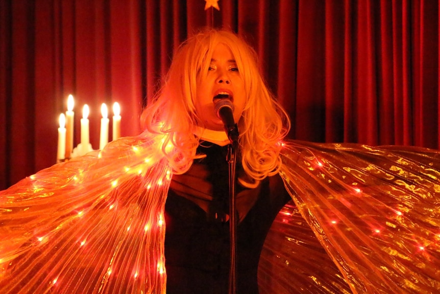 Elly-May wears a wig, sings into a microphone with a light-up cape draped around her.