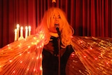Elly-May wears a wig, sings into a microphone with a light-up cape draped around her.