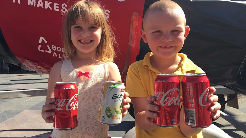 Two children holding empty soft drink can behind a three metre soft drink bottle