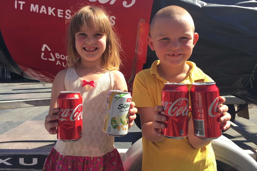 two children holding empty soft drink can behind a three metre soft drink bottle