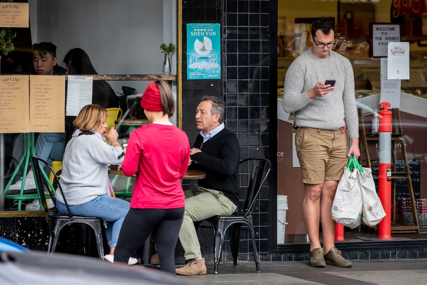 People eat at a table while others stand around them outside a cafe