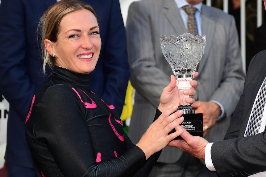 Skye Bogenhuber, wearing pink and black silks, smiles while being handed a crystal trophy.