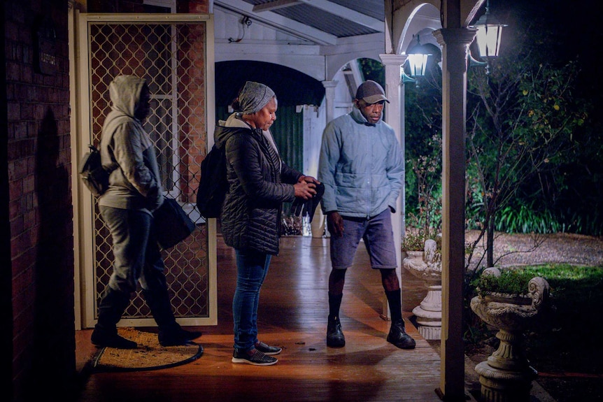 Three workers stand at the front door, it is dark outside and the outdoor lights are on.