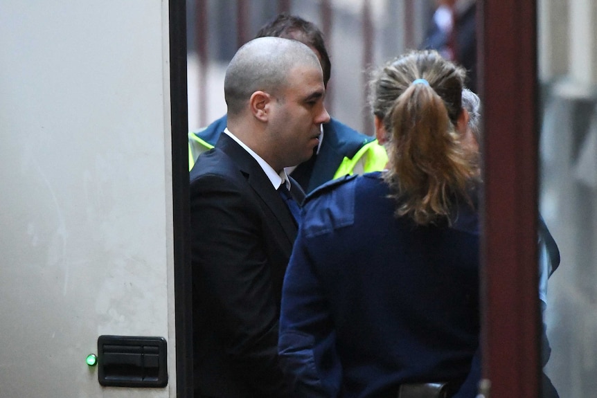 A bald man in a suit is escorted into a building by prison guards.