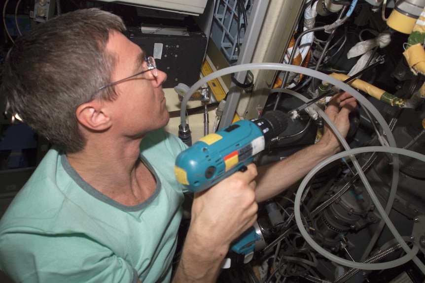 A man in a green T-shirt uses a drill to repair machinery.