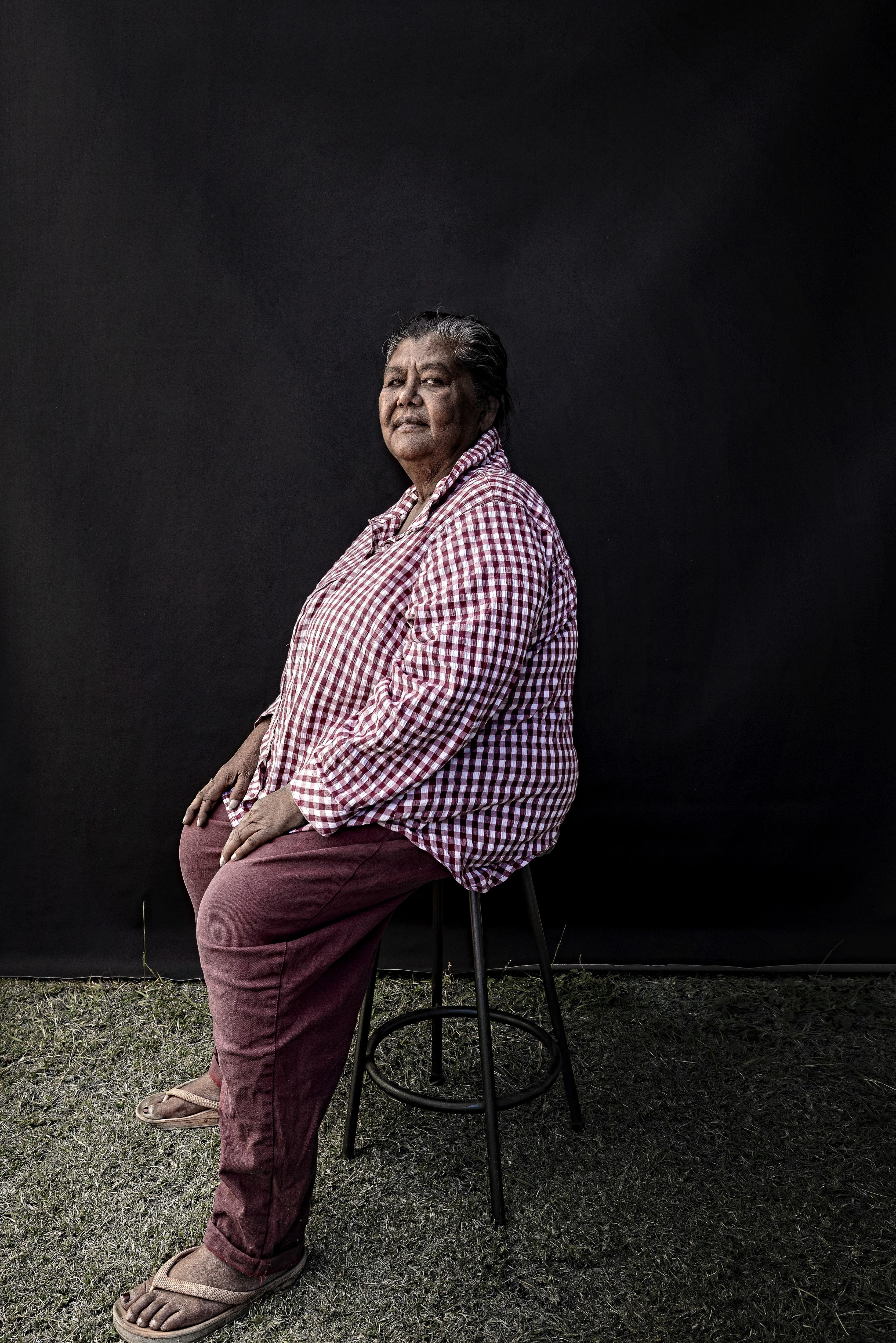 an indigenous woman sits on stool wearing red checkered button up shirt looking into the camera with body turned to the left