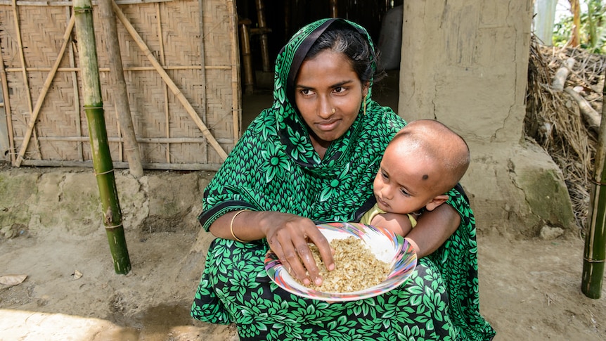 Woman squats with child in arms as she eats a plate of rice outside thatched bamboo home in Bangladesh