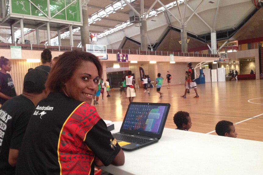 Female sports journalist sits at a desk with a computer on the side of the basketball court