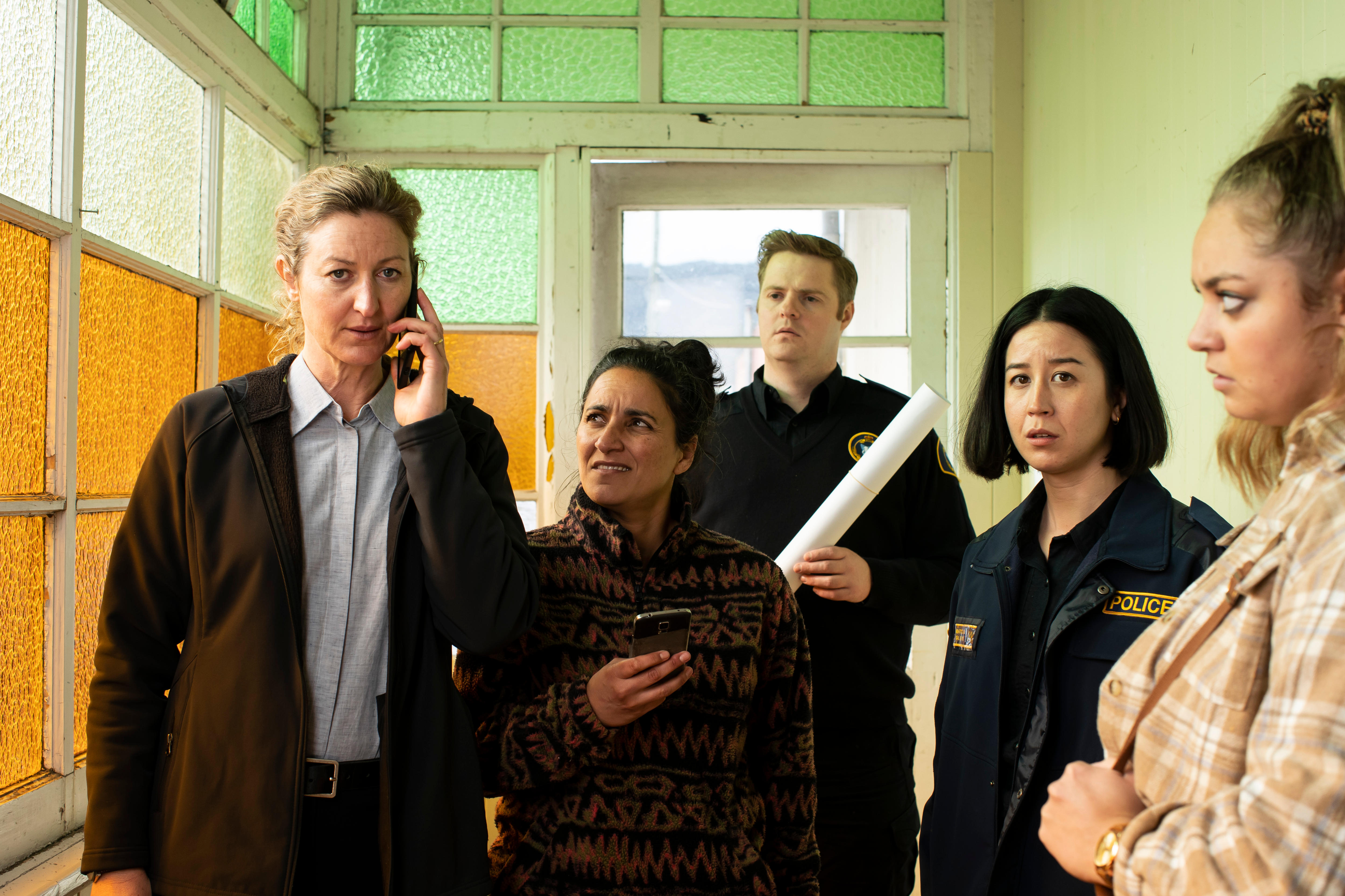 Three women and one man (four are police) stand in a room with stained-glass windows with serious expressions. 