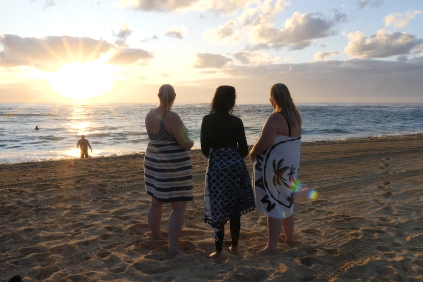 Three women standing together looking at the sunrise at the beach.