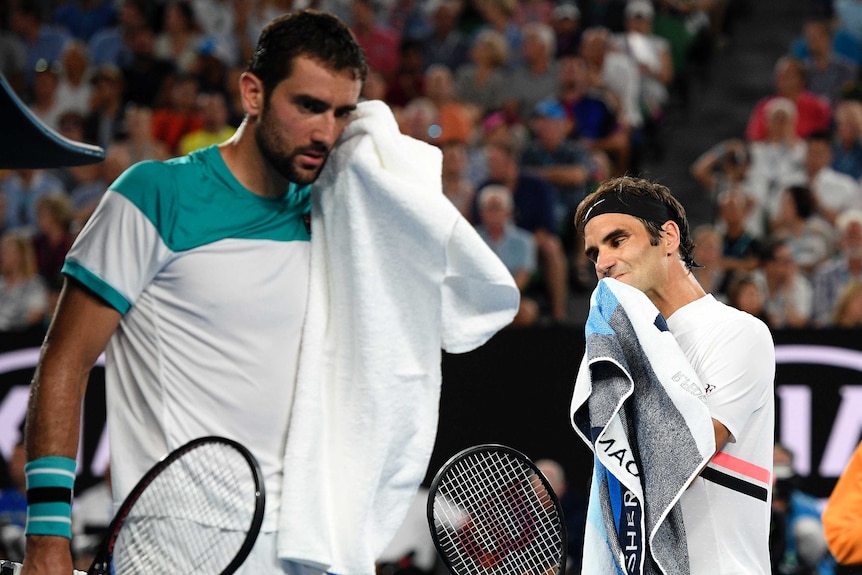 Roger Federer towels off in the background while Marin Cilic does the same in the foreground during a break in a tennis match.