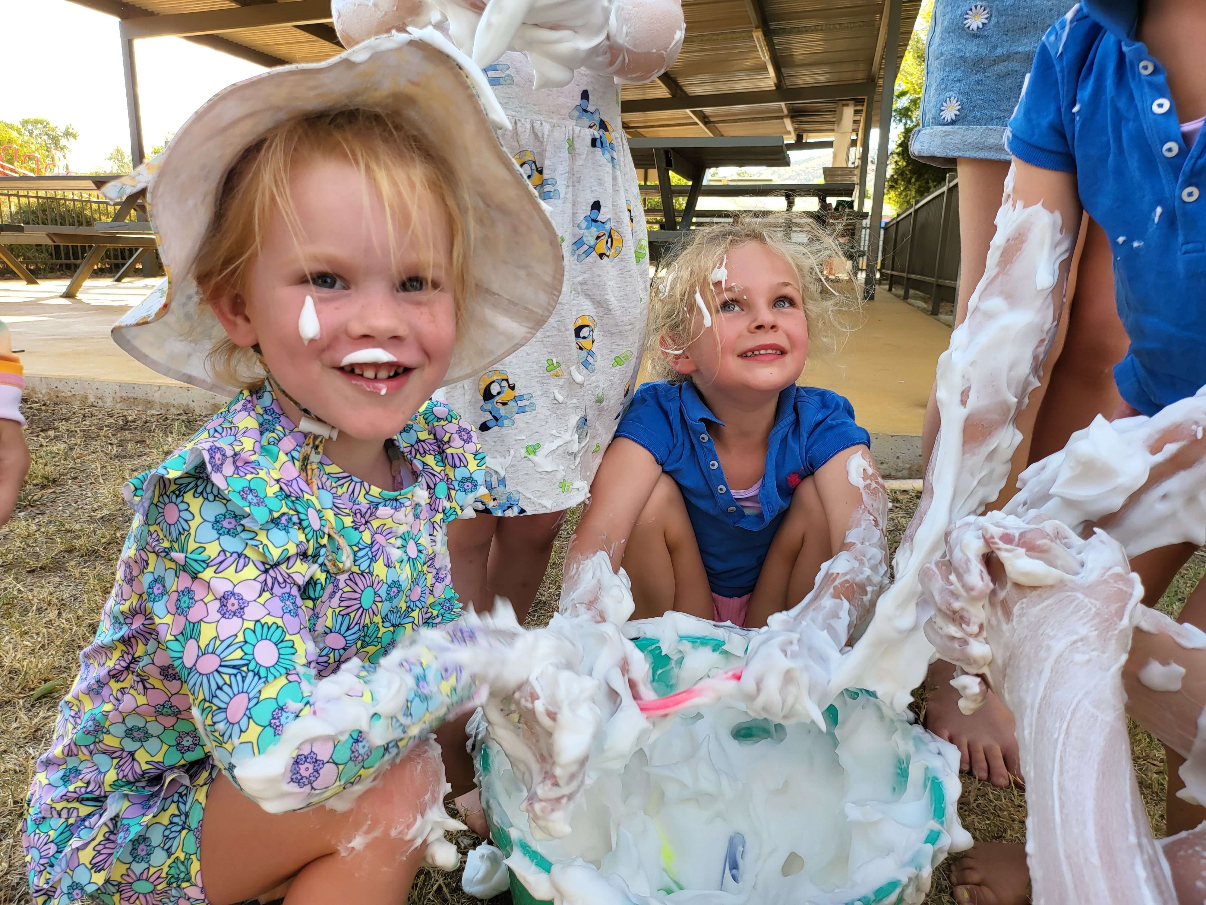 Two little girls smile as they play with a bucket of foam, which has gotten all up their arms