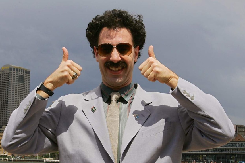Borat in a suit and sunglasses gives a big smile and two thumbs up