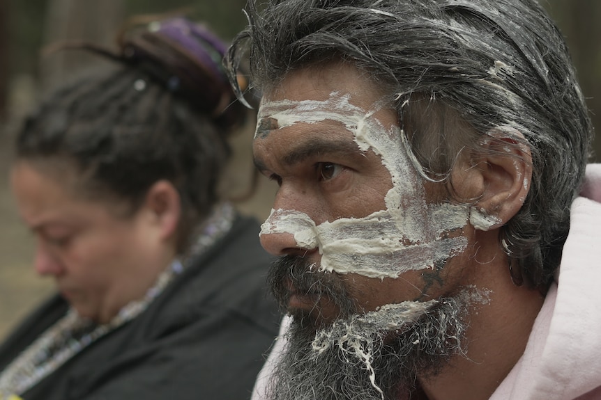 An Aboriginal man sits in profile with white cultural pigment on his face. An Aboriginal woman sits in the background. 