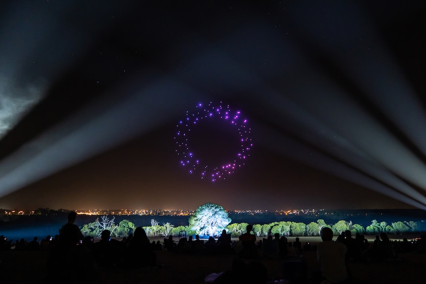 A light show by a lake, including drones, that appear purple, arranged in a circle in the sky, and a spot-lit tree