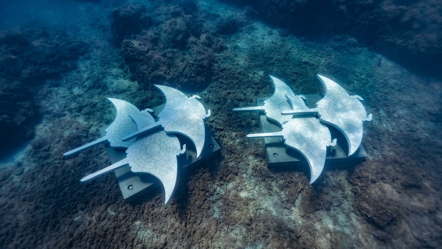 A metal sculpture of two groups of three rays lays on the sea floor.