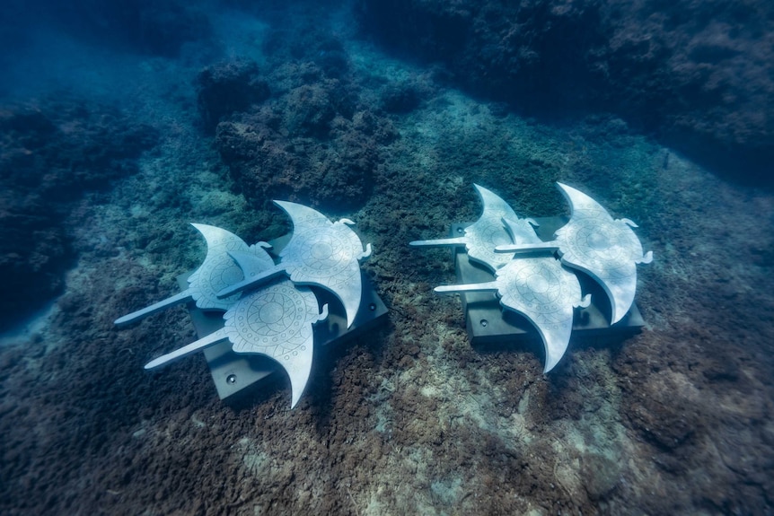 A metal sculpture of two groups of three rays lays on the sea floor.