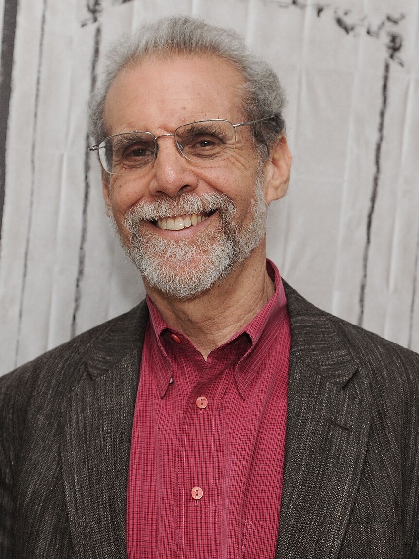 Un homme plus âgé aux cheveux gris, à la barbe grise et aux lunettes sourit à la caméra.