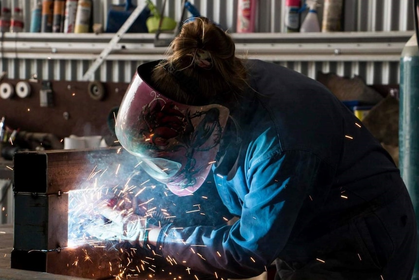 A woman in a pink welding helmet and overalls with sparks flying as she works