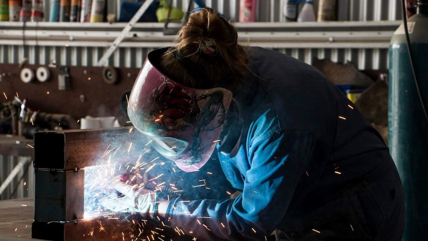 A woman in a pink welding helmet and overalls with sparks flying as she works