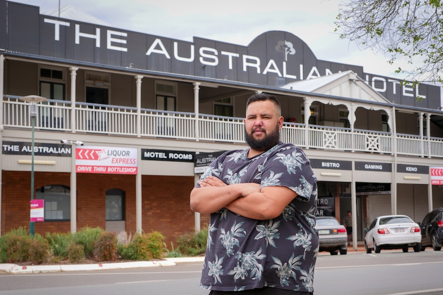 A bearded Indigenous man wearing a floral shirt standing with his arms crossed in front of a large pub.