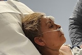 A woman sits in a hospital bed with tubing up her nose and bandages on her face