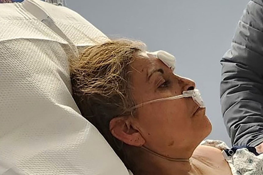 A woman sits in a hospital bed with tubing up her nose and bandages on her face