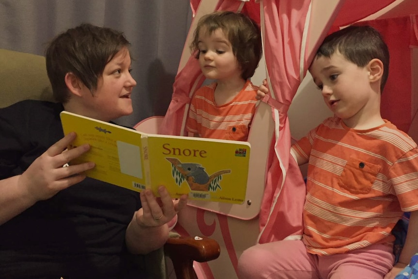 A parent is reading a book to two children who wear matching t-shirts.