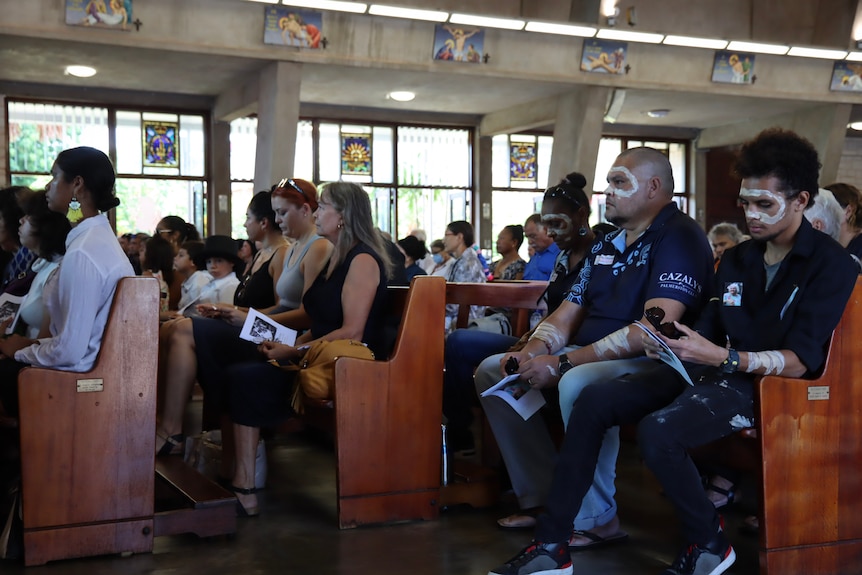 People sitting on pews inside a Catholic church, looking sombre, at a funeral. 